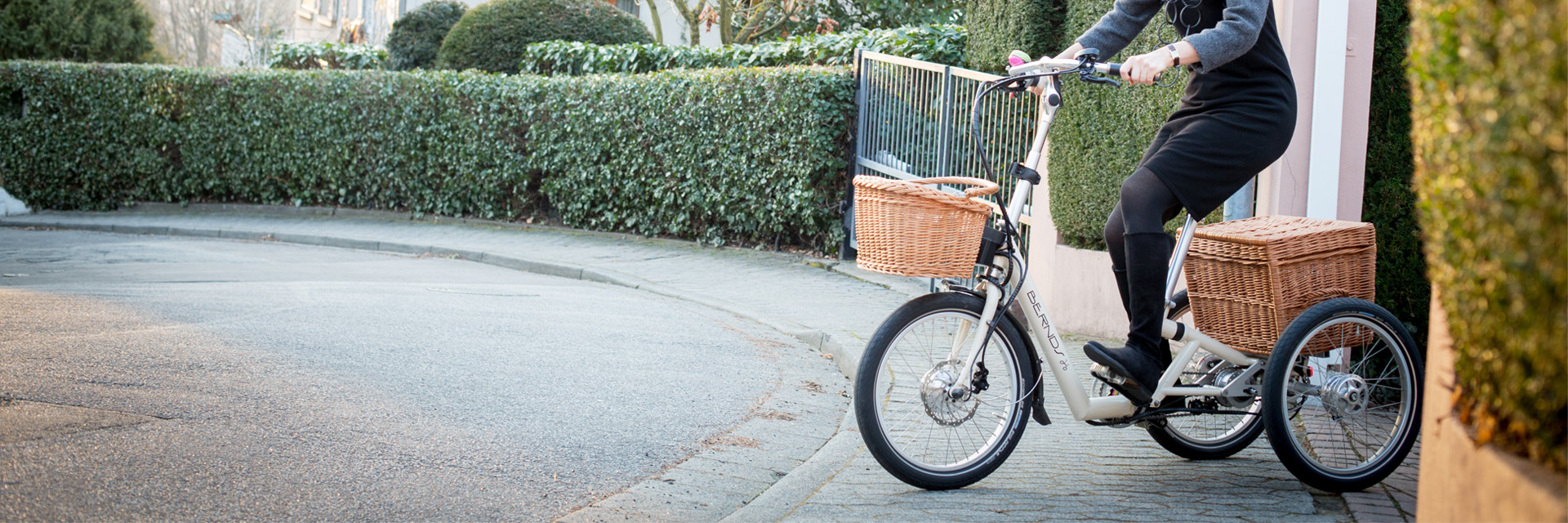 Frau auf dem Bernds Bikes Dreirad Kompakt Grete PickUp auf dem Weg aus einer Einfahrt auf die Straße vor grünen Hecken